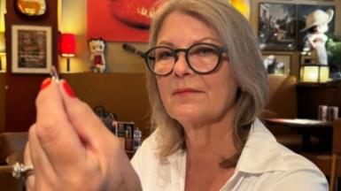 a woman with mid-length grey hair, wearing a white shirt and black-rimmed glasses, holds a shard of glass to the camera. She appears to be in a bar or restaurant 