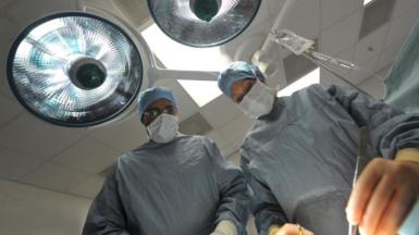 Two surgeons in surgical gear and masks look on in an operating theatre with one of them using a scalpel