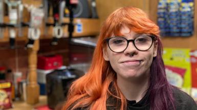 A woman with ginger hair and a purple streak smiles as she looks towards the camera while standing behind a bar. She is wearing glasses with bold black frames, a nose ring and a lip ring. Bottles of spirits and bar equipment can be seen in the background.