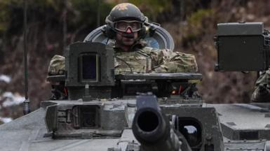 A man in military uniform, tactical goggles and a helmet sits in the control seat of a tank.