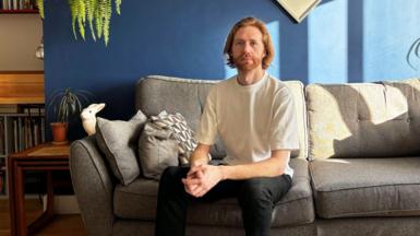 Male sitting on sofa wearing T shirt and dark trousers. It is a home setting with a plant to the left of the sofa. The room is painted blue and the sofa is grey