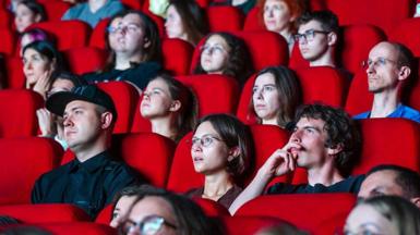 People sit in a cinema auditorium on rows of red chairs.