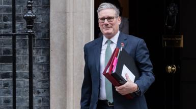 Keir Starmer leaves Downing Street while holding folders and wearing glasses and a suit and smiling slightly on Wednesday