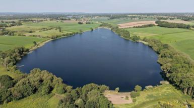 Aerial shot of Pickmere Lake in Cheshire on a sunny day, a wide body of water surrounded by green spaces.