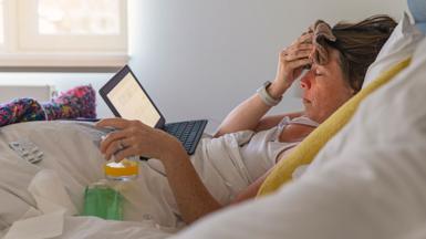 Woman in bed with the flu or a cold she is has an orange drink and has a flannel on her head. She is looking at a laptop