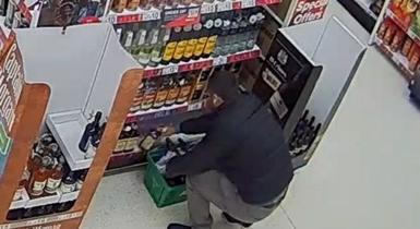 Man crouching down on a supermarket floor, next to shelves of alcohol bottles. He is putting them into a green shopping basket.