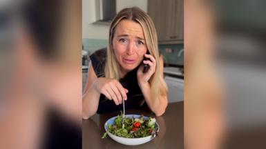 Person sitting at a kitchen table holding a mobile phone to one ear while eating salad