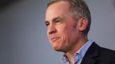 Mark Carney, in an open collared blue shirt and suit jacket, with a slight smile. 