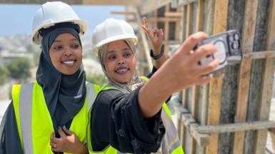 Saadia Ahmed Omar (right) takes a photo of herself and Fathi Mohamed Abdi (left) atop a building under construction in Mogadishu. They are both wearing hard hats over their headscarves and are in high vis yellow vests. Ms Omar makes the victory sign as she takes the photo.
