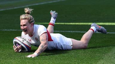 Meg Jones, wearing a white England shirt, shorts, and socks with her blonde hair tied up in a bin, grins as she dives over the tryline with the ball in her right hand