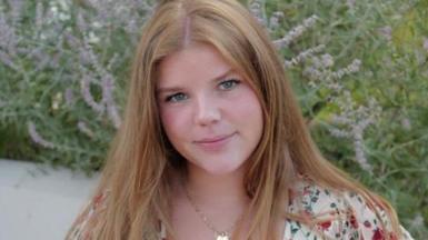 Orla Wates, a girl with long, ginger hair, smiles at camera. She is wearing a white top with red flowers and sits in front of a flowering lavender plant.