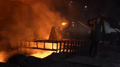 A steelworker at Blast Furnace 4 tests the liquid iron that glows orange as it emerges from the furnace