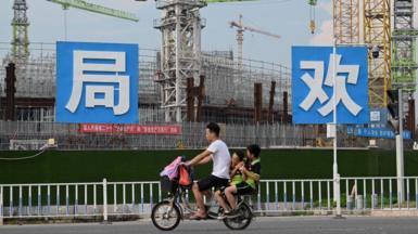 People commute in front of the under-construction Guangzhou Evergrande football stadium in Guangzhou, China's southern Guangdong province on September 17, 2021. The photo shows a male motorcyclist with two passengers, children, seated behind him, as they ride past a large construction site.