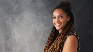 A woman with long braided afro hair is smiling as she is standing next to a grey backdrop.