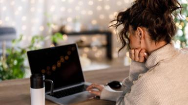 A stock photo shows a young woman sat at a wooden table with an open laptop, resting their chin on one hand. A reusable travel mug sits nearby.