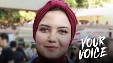 Susan Nasser, a young woman wearing a burgundy turban, pictured outside with people in the background. The words YOUR VOICE have been imposed on the photo on the right hand side.