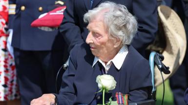 Christian Lamb, a 103-year-old woman with white hair wearing a navy blue jacket and white shirt sits outside in a wheelchair and holds a white rose