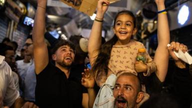 People cheer and hold placards as they celebrate the news of the death of Hamas leader Yahya Sinwar, in Jerusalem. A man has a smiling young girl on his shoulders. People behind him wave smartphones and signs in the air.