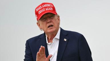 A close up shot of President Donald Trump speaking to outside with his hand raised. He is wearing a "make America great again" cap