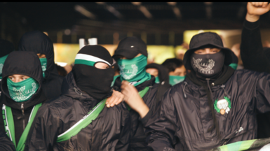 A group of individuals dressed in black jackets and green-and-black scarves or masks stand closely together in a crowded setting, with raised arms and coordinated clothing visible under dim lighting.
