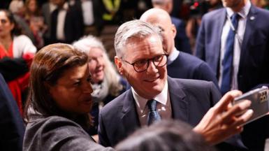 Sir Keir Starmer smiles and poses for a selfie with an admirer, he is dressed in a suit and tie and is wearing glasses at an event at Pinewood studios