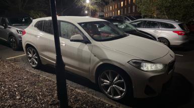 White BWM saloon car with lights on parked in a residential parking area with serveral other vehicles two are silver. Parked up near a streetlight with a two story brick building in the background. There is an area covered in bark chippings next to the white car.