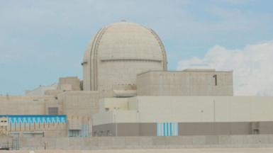 A large sand-coloured building in the UAE called the Barakah Nuclear Power Plant. In the centre is a large dome while rectangular shaped sections of the building lie to the left, right, and in front of the dome. The sky behind is blue with a few white clouds.