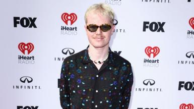 Lloyd Wakefield stands in front of a white backdrop that has Fox, iHeart Radio and Infiniti logos. He has short light blonde hair, a moustache and is wearing sunglasses. He is smiling at the camera and is wearing a black, floral beaded shirt with a colourful charm necklace underneath.