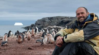 Derren Fox is sitting on the ground smiling at the camera. He has a beard and moustache and is wearing a black and yellow anorak, jeans and walking boots. He is surrounded by dozens of penguins and there is a cliff and the sea behind him. Large icebergs are in the sea