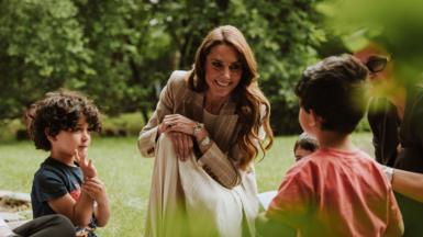 A smiling Kate wearing a brown blazer and a long white dress crouches with several small children in a garden