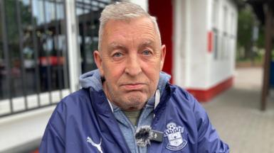 A man with short grey hair looks upset sat outside a red and white building. He is wearing a blue Southampton FC jacket and a grey hoodie.