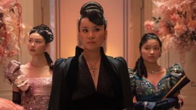 Araminta Gun stands in the centre of a pink floral hall entrance wearing a wavey hair updo, black dress and silver necklace. She is between her two daughters, one of whom is in a pink sparkling dress, the second is in a blue sparkling puff sleeve dress and wears navy blue gloves