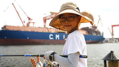 A young man is smiling at the camera as he fishes on the dock. He is weating a sun hat, spectacles and a white t-shirt over a full-sleeved shirt. He has a watch on his left hand, wihich is holding the black fishing rod. 

Behind him is a long blue ship, with the letters C. M L on it in white. There are large cranes hovering over the vessel.     