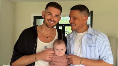 Adam Frisby, who is wearing a black jacket and white T-shirt, and Jamie Corbett, who is wearing a blue striped shirt with a white top underneath, are standing in their living room and holding their baby girl Leven. 
