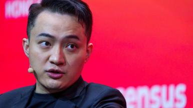 Justin Sun in black blazer and black T-shirt, with small beige microphone and ear piece in front of red background