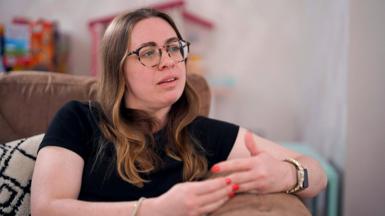 Katy, who wears glasses and a black T-shirt, talks to BBC Panorama from her sofa. Toys can be seen in the background.