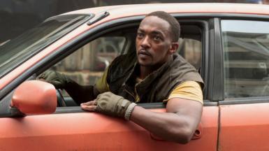 A black man with short hair and a neat moustache leans out of the open window of a red-orange car. He wears a cargo jacket with the arms cut off over a yellow t-shirt and fingerless gloves. He's got one hand on the steering wheel and the other arm resting on the car's door. He's looking at someone or something out of shot and appears to be listening attentively or staring with trepidation.