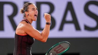 Aryna Sabalenka, wearing a black dress and holding her green racquet in her left hand, screams and clenches her right fist in celebration