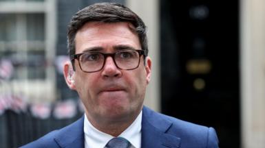 Close-up of Andy Burnham wearing tortoiseshell glasses, a light blue suit and tie and white shirt, standing in front of a blurred No 10 Downing Street