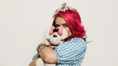 Woman in blue and white checked shirt with dyed pink hair and a tartan hair bow hugging white teddy.