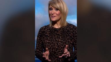 Young woman with blonde hair and patterned dress holding her hands in front of her.