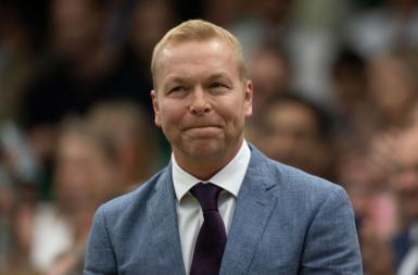 Sir Chris Hoy standing to receive the crowd at Wimbledon in 2024. He looks calm and with a neutral experience, wearing a light blue tweed suit and a purple tie with a white shirt