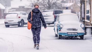 A woman walks down a snowy street in a winter coat and neck warmer, while carrying an orange shoulder bag, and with cars and buildings in the background covered in snow, in Goathland in Yorkshire on Wednesday