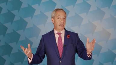 Nigel Farage talking at a conference. He has grey hair and is wearing a blue suit with a red patterned tie. 