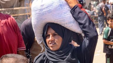 A Palestinian woman carries a bag of food on her head in Rafah, Gaza. She is wearing a black outfit and black headscarf. Behind her is a boy and other people.