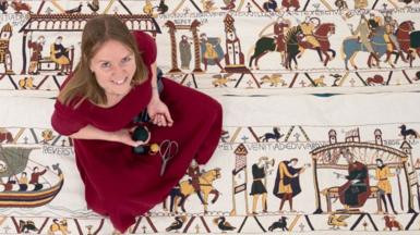 Mia Hansson, wearing a maroon dress, sits on her replica of the Bayeux Tapestry, looking up at the camera and smiling. On her lap is some thread and a pair of scissors.