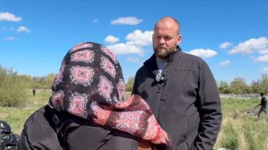 James Waterhouse in a black jacket speaking to a woman in a gilet and pink and black headscarf