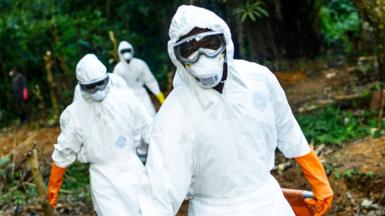 The image depicts a group of individuals dressed in full-body protective suits, complete with face masks, goggles, and bright orange gloves. Their attire suggests they are engaged in a high-risk operation, likely involving hazardous materials or infectious disease control. They are working outdoors in what appears to be a forested or rural environment, with greenery and natural terrain surrounding them. The scene conveys a sense of urgency and seriousness, as the protective gear is designed to minimise exposure to dangerous contaminants.