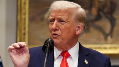 Close-up picture of Donald Trump with his right hand in the air. He is speaking with a painting in the background blurred. He is wearing a blue suit, white shirt and red tie.