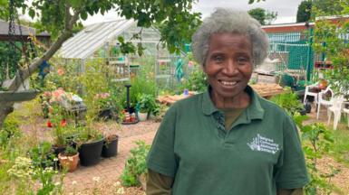 A woman in a green T-shirt which says Belgrave Community Allotment and Garden on it stands in a green space with pot plants, greenhouses, plant beds and white plastic chairs. She has white short hair and is smiling.